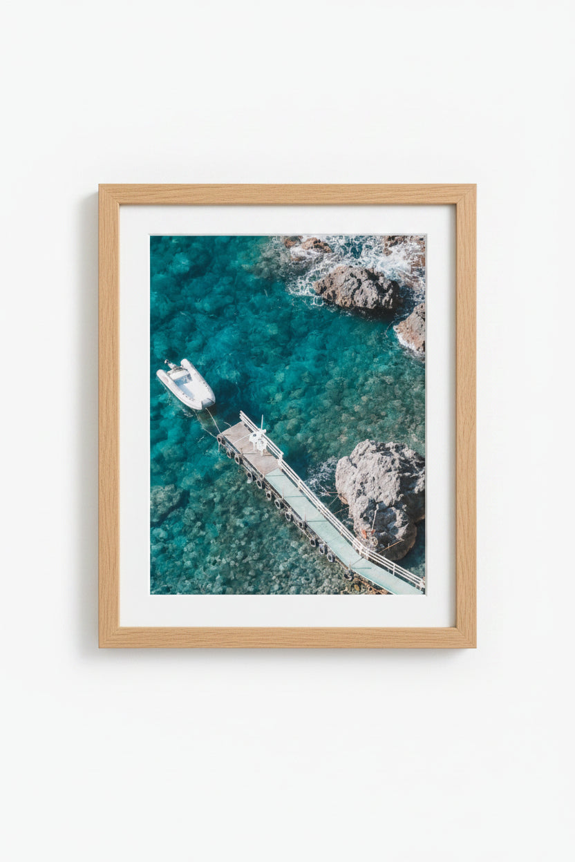 A framed print featuring a photograph by Brandon Hirsler, with a view of a boat docked at a pier in blue waters, surrounded by rocks. Amalfi Beach Scape 2 — Brandon Hirsler Framed Print