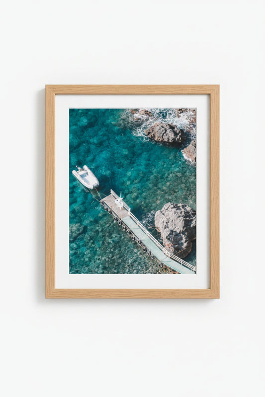A framed print featuring a photograph by Brandon Hirsler, with a view of a boat docked at a pier in blue waters, surrounded by rocks. Amalfi Beach Scape 2 — Brandon Hirsler Framed Print